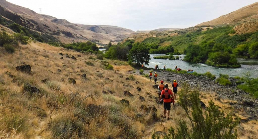 a group of students walk through a grassy and rocky shore, toward a blue river